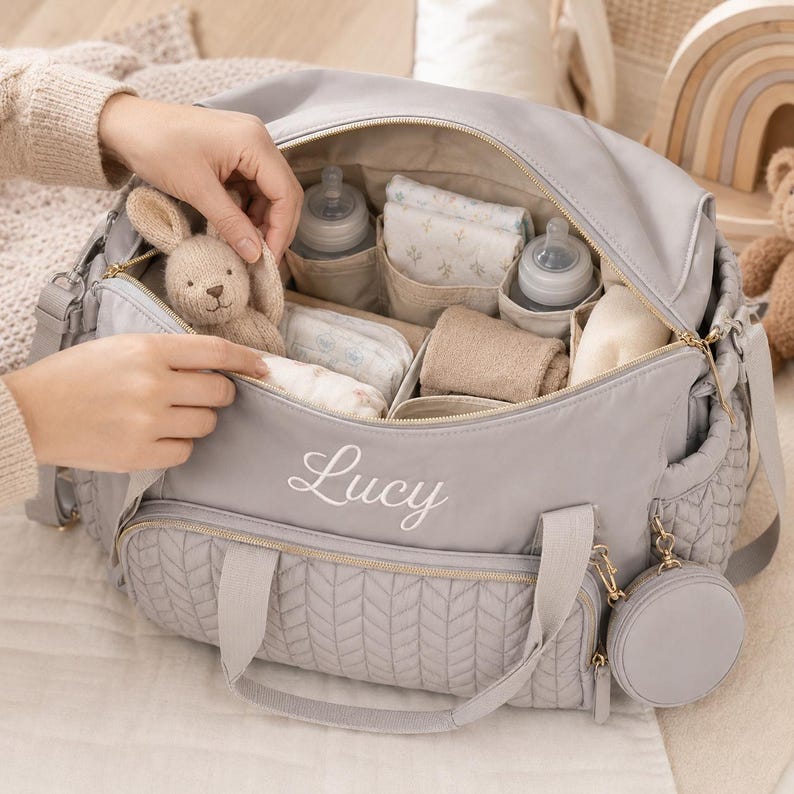 May include: A light gray diaper bag with a quilted front pocket and the name Lucy embroidered in white script. The open bag contains two baby bottles, folded cloths, and a small knitted rabbit toy. A matching circular pouch hangs from the side.