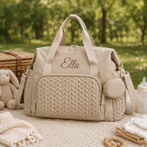May include: A beige diaper bag featuring a quilted herringbone pattern on the front pocket and side panels. The bag has two top handles, a detachable shoulder strap, and a small circular pouch. The name Ella is embroidered in dark brown script.