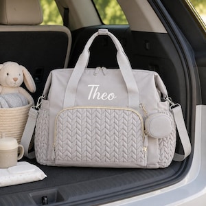 May include: A light gray diaper bag featuring a quilted front pocket with a chevron pattern, two top handles, and a detachable shoulder strap. The name Theo is embroidered in white script on the front. A small matching circular pouch is attached.