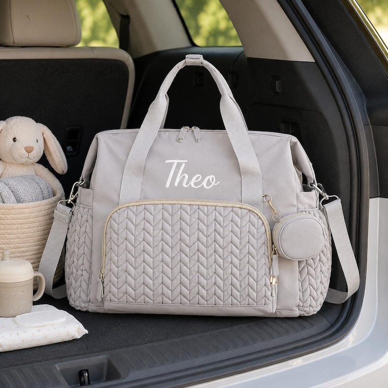 May include: A light gray diaper bag featuring a quilted front pocket with a chevron pattern, two top handles, and a detachable shoulder strap. The name Theo is embroidered in white script on the front. A small matching circular pouch is attached.