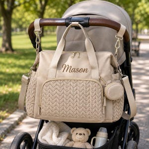 May include: A beige diaper bag with a quilted chevron pattern on the front pocket and side panels. The bag features the name Mason embroidered in brown thread, gold-tone zippers, two top handles, a detachable shoulder strap, and a small round pouch.