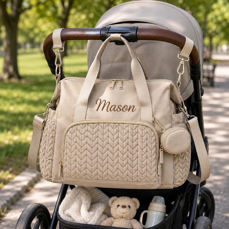 May include: A beige diaper bag with a quilted chevron pattern on the front pocket and side panels. The bag features the name Mason embroidered in brown thread, gold-tone zippers, two top handles, a detachable shoulder strap, and a small round pouch.