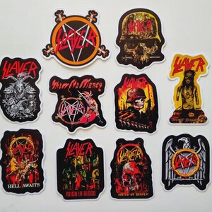 May include: A collection of Slayer band stickers in various designs. The stickers feature the band's logo, skulls, and other imagery in red, black, and white. Some stickers include text such as "Hell Awaits" and "Reign in Blood".