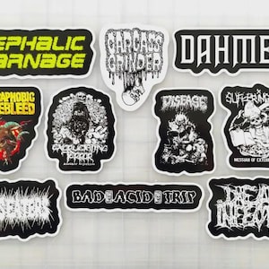 May include: A collection of black and white stickers with various band names and graphic designs. The stickers include text such as "Cephalic Carnage", "Carcass Grinder", and "Dahmer". Other stickers feature illustrations of skulls and grotesque figures.