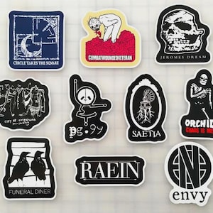 Pode incluir: Uma coleção de autocolantes com vários designs e texto. Os autocolantes apresentam ilustrações de uma caveira, um esqueleto, um panda e outros gráficos. O texto inclui "CIRCLE TAKES THE SQUARE", "COMBATWOUNDEDVETERAN" e "FUNERAL DINER".
