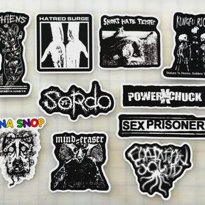 May include: A collection of black and white stickers with various designs and text. The stickers feature phrases like "CHIENS," "HATRED SURGE," and "KUNGFU RICK." Some stickers have illustrations, including a hand, a figure, and a creature. The stickers are on a white surface.