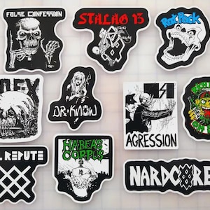 May include: A collection of punk rock stickers with various designs. The stickers feature skulls, band logos, and graphic illustrations in black, white, red, and green. Text includes "FALSE CONFESSION", "STALAG 13", "Rat Pack", "NOFX", "DR. KNOW", "AGGRESSION", "ILL REPUTE", "HABEAS CORPUS", and "HARDCORE".