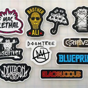 May include: A collection of stickers with various designs and text. The stickers feature phrases like "MAC LETHAL," "BROTHER ALI," and "BLUEPRINT." The color palette includes black, white, red, and blue, with some stickers resembling a skateboard.