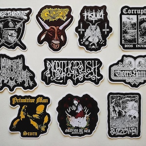 May include: A collection of black and white stickers featuring various band logos and artwork. Designs include a pig with a helmet, a horned demon, a skeletal figure, and the text "Corrupted". These stickers are likely for fans of heavy metal music.