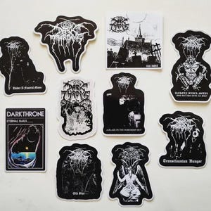 Puede incluir: Una colección de pegatinas en blanco y negro con logotipos de bandas góticas y de metal y arte de álbumes. Las pegatinas incluyen imágenes de paisajes, figuras y texto. Las pegatinas están dispuestas sobre una superficie blanca.