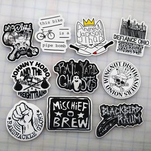 May include: A collection of black and white stickers with various designs. The stickers feature text and illustrations, including a pig with a guitar, a bicycle with the text "pipe bomb", a cat with knives, and a cityscape. Other stickers include a fist, a feather, and a skull.