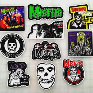 Puede incluir: Una colección de pegatinas de la banda Misfits con varios diseños. Las pegatinas muestran el logotipo de la banda, imágenes de calaveras y portadas de álbumes, con colores como verde, rojo, negro y blanco. Algunas pegatinas incluyen texto como "Fiend Club" y "Die Die My Darling."