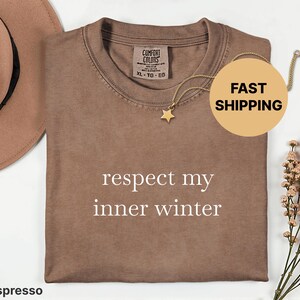 May include: Espresso-coloured t-shirt with the text "respect my inner winter" in white. A gold star pendant necklace and a brown hat are next to the shirt. The shirt has a Comfort Colors tag.