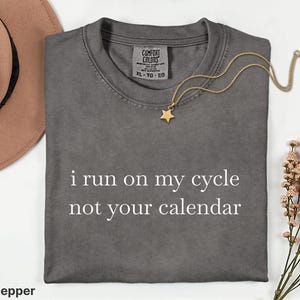 Puede incluir: Una camiseta gris carbón de manga corta con el texto blanco "i run on my cycle not your calendar" impreso en el frente. La camiseta cuenta con una etiqueta de la marca Comfort Colors en el cuello interior, talla XL, y es 100% algodón.