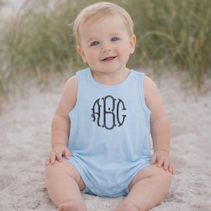 May include: A light blue baby romper with a dark gray monogram design. The sleeveless romper features a round neckline and snap closures. The monogram includes the letters "ABC" in an elegant, stylized font. The baby is sitting on a sandy surface.