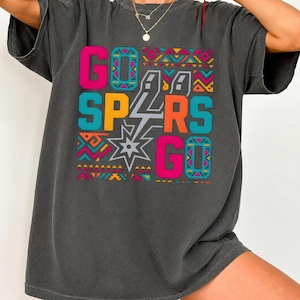 Puede incluir: Camiseta gris oscuro con las palabras "GO SPURS GO" en rosa brillante, naranja, turquesa y amarillo. El texto est&aacute; sobre un patr&oacute;n geom&eacute;trico con una estrella y un rayo. Corte holgado y casual.