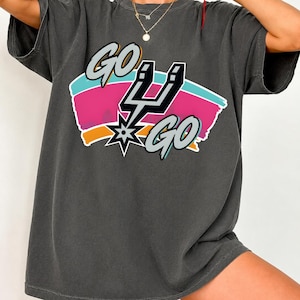 May include: Dark grey t-shirt with the text "GO GO" in a stylized font, accompanied by a lightning bolt and star graphic. The design incorporates pink, blue, and orange colours. The shirt has short sleeves and a relaxed fit.