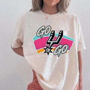 May include: Cream-coloured t-shirt with the words "GO SPURS GO" in a graphic design. The design includes a black and silver Spurs logo, set against pink, blue, and orange stripes. The shirt has short sleeves.