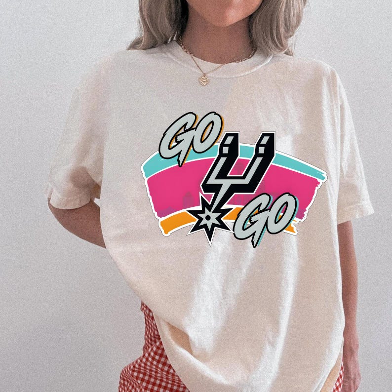 May include: Cream-coloured t-shirt with the words "GO SPURS GO" in a graphic design. The design includes a black and silver Spurs logo, set against pink, blue, and orange stripes. The shirt has short sleeves.