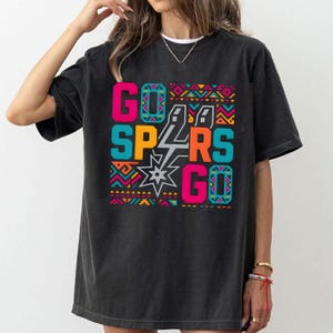 Puede incluir: Camiseta negra con un dise&ntilde;o gr&aacute;fico colorido. El dise&ntilde;o presenta las palabras "GO SPURS GO" en rosa brillante, turquesa y naranja, con una estrella y el logotipo de los Spurs. El dise&ntilde;o est&aacute; enmarcado por un patr&oacute;n geom&eacute;trico.