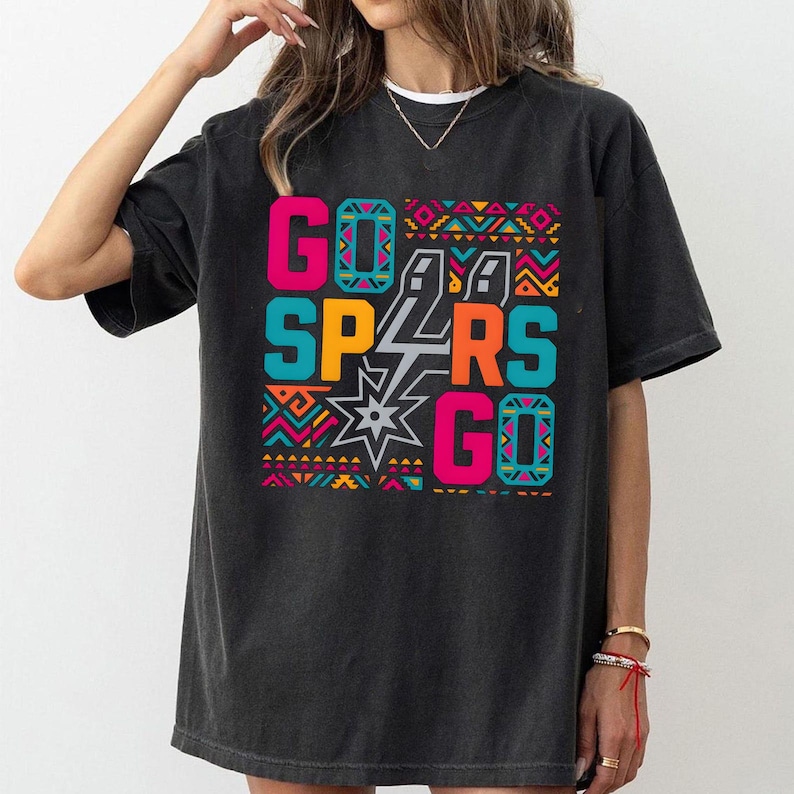 Puede incluir: Camiseta negra con un dise&ntilde;o gr&aacute;fico colorido. El dise&ntilde;o presenta las palabras "GO SPURS GO" en rosa brillante, turquesa y naranja, con una estrella y el logotipo de los Spurs. El dise&ntilde;o est&aacute; enmarcado por un patr&oacute;n geom&eacute;trico.