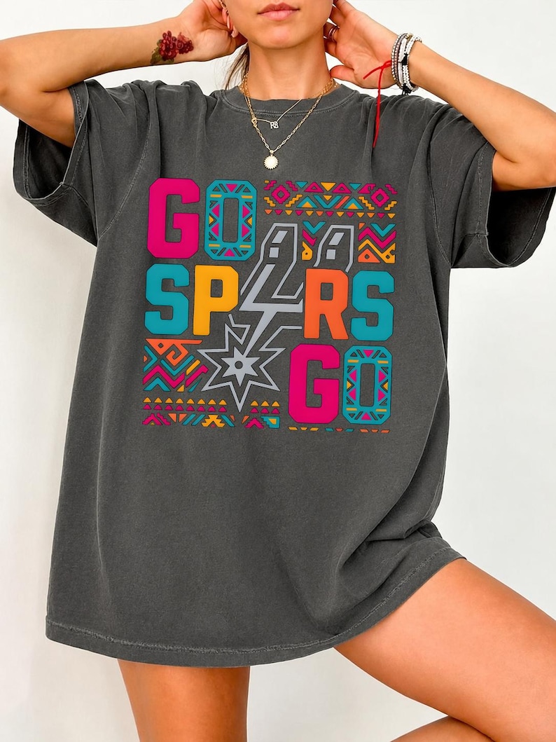 Puede incluir: Camiseta gris oscuro con las palabras "GO SPURS GO" en rosa brillante, naranja, turquesa y amarillo. El texto est&aacute; sobre un patr&oacute;n geom&eacute;trico con una estrella y un rayo. Corte holgado y casual.
