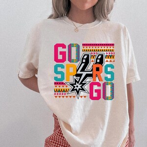 Puede incluir: Camiseta color crema con las palabras "GO SPURS GO" en un dise&ntilde;o geom&eacute;trico colorido. El dise&ntilde;o incluye el logotipo de los Spurs y varios patrones en rosa, turquesa, naranja y negro.