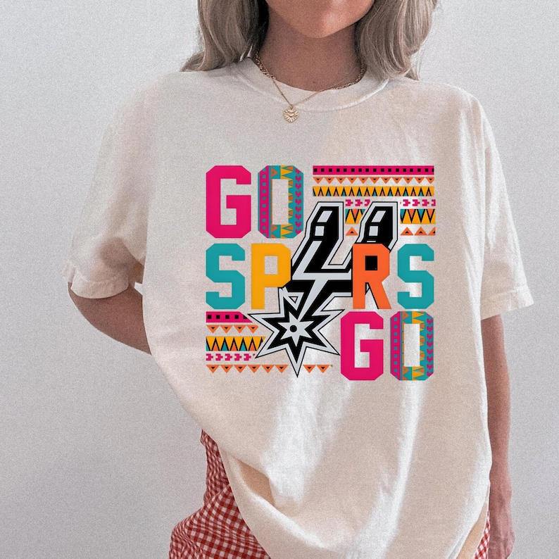 Puede incluir: Camiseta color crema con las palabras "GO SPURS GO" en un dise&ntilde;o geom&eacute;trico colorido. El dise&ntilde;o incluye el logotipo de los Spurs y varios patrones en rosa, turquesa, naranja y negro.