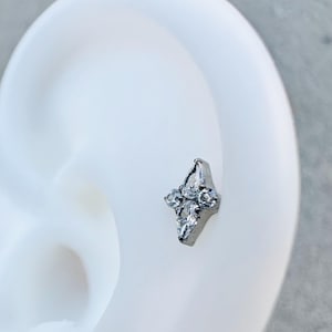 Op de afbeelding: Een zilveren oorbel met een geometrisch ontwerp met heldere edelstenen. De oorbel heeft de vorm van een gestileerde bloem of sterrenuitbarsting, met een centraal punt en meerdere omliggende stenen. De sieraden worden op een witte achtergrond getoond.