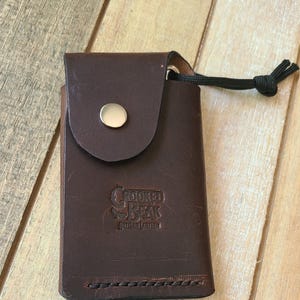 May include: A dark brown leather wallet with a flap closure secured by a gold-colored snap. The wallet has a black cord attached and the embossed text "Crooked Beak Artisan Leather". The wallet is rectangular and appears to be new.