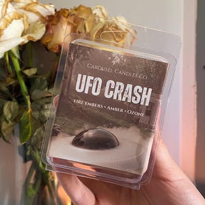 Può includere: Una confezione di plastica trasparente contenente una barra di cera profumata. L'etichetta recita "UFO CRASH" con il logo Carousel Candles Co. Le note olfattive sono "Fire Embers, Amber, Ozone". L'immagine sull'etichetta raffigura un UFO.