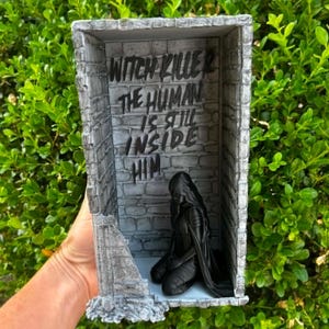May include: A gray, rectangular diorama featuring a stone-textured wall with the black text "WITCH KILLER THE HUMAN IS STILL INSIDE HIM" printed on it. A black, kneeling figure is positioned on the floor of the structure, which has open sides.