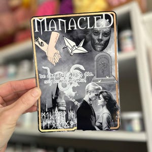 May include: A black and white sticker with a collage of images, including a couple, a castle, a snake, a tombstone, a paper crane, and a mask. The text "MANACLED" is at the top of the sticker. The text "be careful, don't die. only because you asked." is in the middle of the sticker.