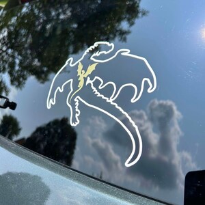 May include: A white vinyl sticker of a dragon silhouette on a dark green car window. The dragon is flying with its wings spread and its tail curled.