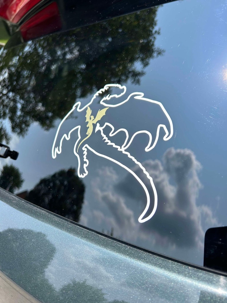 May include: A white vinyl sticker of a dragon silhouette on a dark green car window. The dragon is flying with its wings spread and its tail curled.