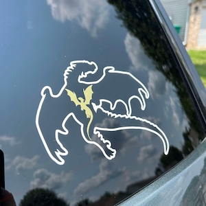 May include: A white silhouette of a dragon with a smaller golden dragon inside, on a black car window.
