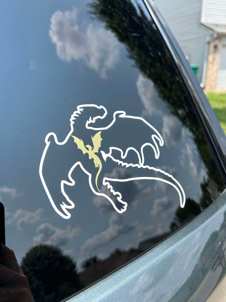 May include: A white silhouette of a dragon with a smaller golden dragon inside, on a black car window.