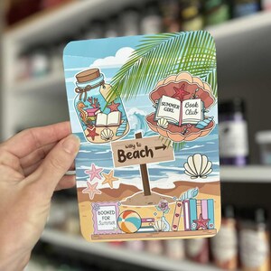 May include: A colorful beach-themed card featuring illustrations of a jar with sea creatures, an open book, and a seashell with text that reads "Summer Girl Book Club." The card also includes a sign that says "Way to Beach" and the text "Booked for Summer."