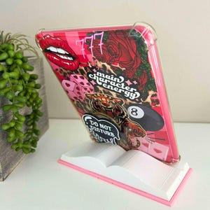 May include: A pink tablet case with a bold design featuring red lips, roses, dice, a tiger, and the text "main character energy" and "Do Not Disturb". The case is displayed on a white book-shaped stand with pink edges.
