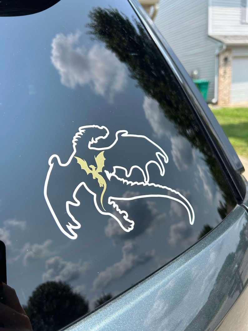 May include: A white vinyl sticker of a dragon silhouette with a smaller dragon silhouette inside. The sticker is on a black car window.