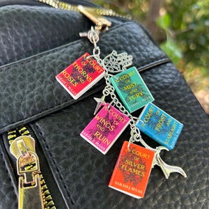May include: A silver charm featuring miniature book pendants with titles like "A Court of Thorns and Roses" and "A Court of Mist and Fury." The charm is attached to a black bag with a gold zipper.