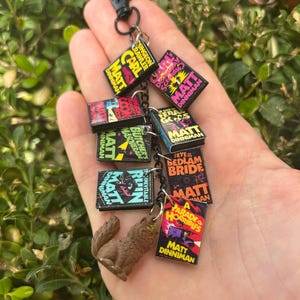 May include: A keychain featuring miniature book charms and a small wolf figurine. The book charms have colorful covers with the name "Matt Dinniman" and various titles. The keychain is attached to a black clasp.