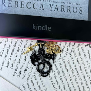 May include: A black dragon pendant with gold accents and a lightning bolt charm hangs over a Kindle e-reader. The e-reader is on top of a book with text from the author Rebecca Yarros.