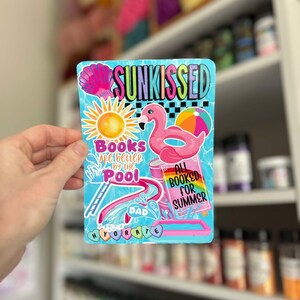 May include: A blue and pink sticker with the text "SUNKISSED" in rainbow letters. The sticker features a pink flamingo pool float, a sun, a book with the text "ALL BOOKED FOR SUMMER", a pool slide, and the text "Books are better by the pool" and "HYDRATE".