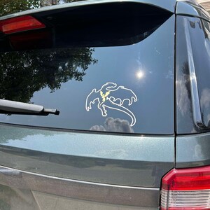 May include: A dark green SUV with a dragon silhouette sticker on the rear window. The sticker is white with a yellow lightning bolt design.