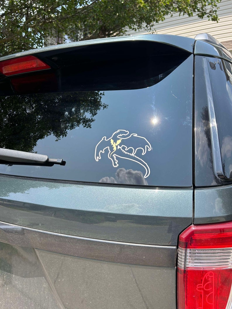 May include: A dark green SUV with a dragon silhouette sticker on the rear window. The sticker is white with a yellow lightning bolt design.