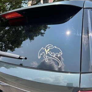 May include: A green car with a white dragon sticker on the back window. The dragon is facing to the right and has a yellow lightning bolt on its chest.