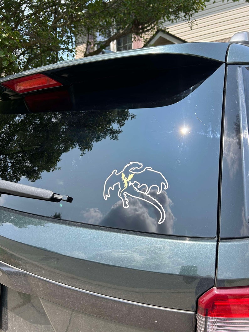 May include: A green car with a white dragon sticker on the back window. The dragon is facing to the right and has a yellow lightning bolt on its chest.