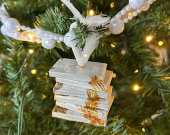 May include: A stack of miniature books with gold accents and text, hanging as a Christmas tree ornament. The books are white with gold lettering and a decorative design. The ornament is suspended from a white beaded garland on a green Christmas tree.