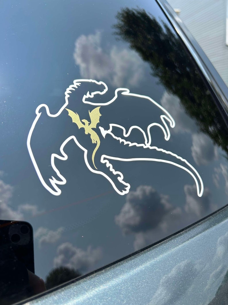 May include: A white silhouette of a dragon with a smaller gold dragon inside, on a black car window.